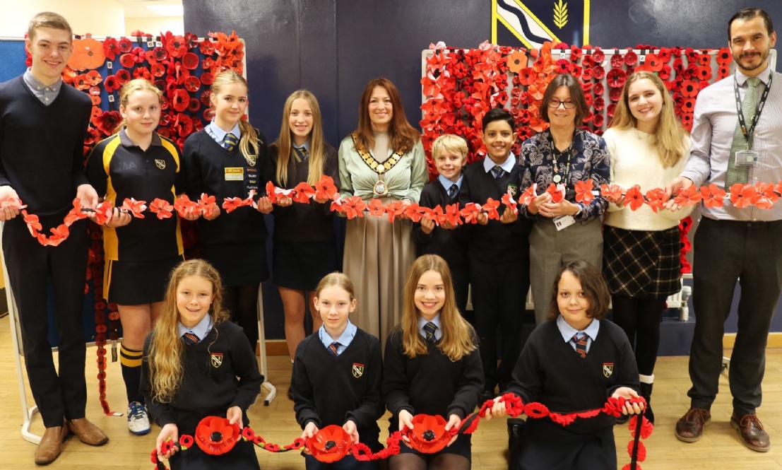 Group of school pupils and teachers holding a chain of red paper poppies in front of poppy displays on the wall, commemorating Remembrance Day.